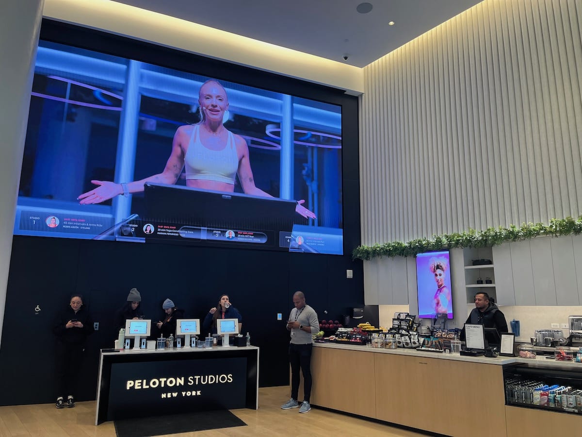 A fitness instructor appears on a large screen in an indoor space with a "peloton studios new york" information desk in the foreground.